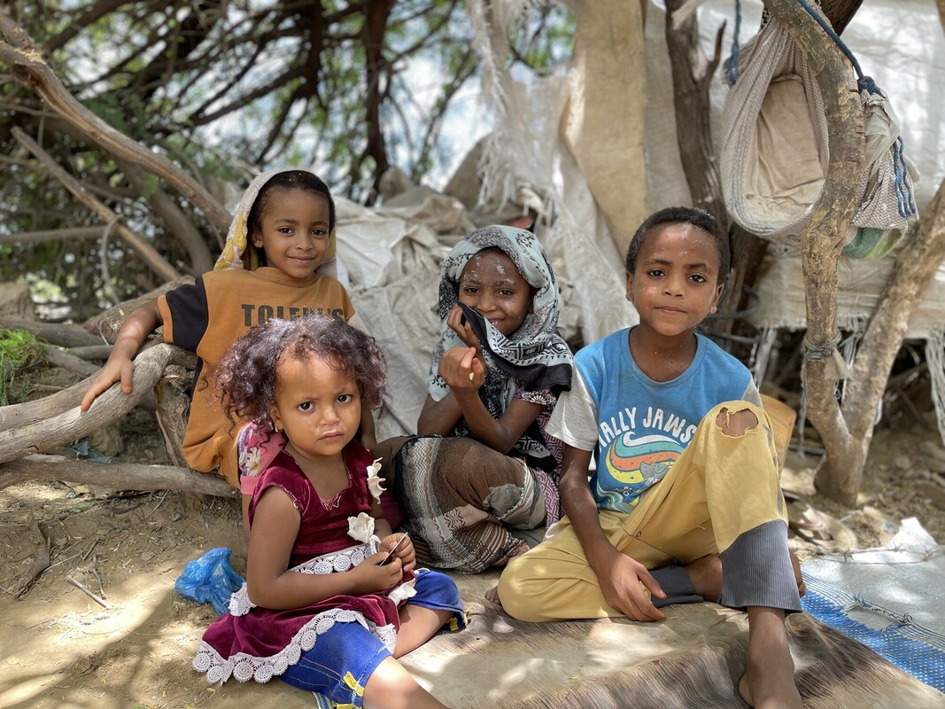 Four smiling Yemeni children sitting on the ground under a tree.